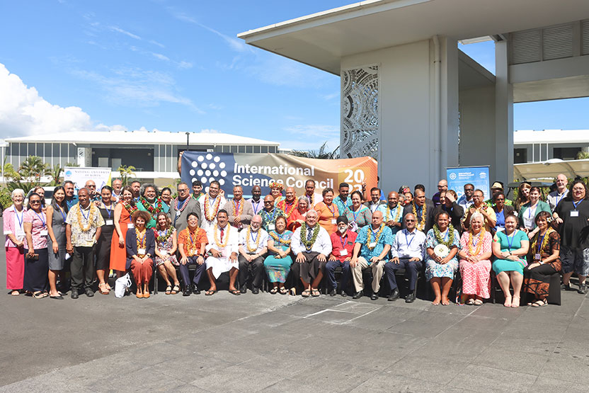A large group of people in colourful clothes and tropical flower leis gather for a group photo in front of a banner that reads ‘International Science Council’