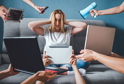 A woman sits on a couch surrounded by hands proffering laptops, phones, tablets and a megaphone. She appears overwhelmed and has her hands covering her ears.