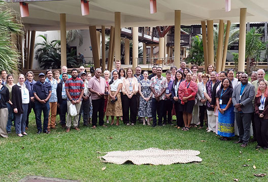A large group of people posing for a group photo on a grassy area in front of a building, with a woven mat in the foreground