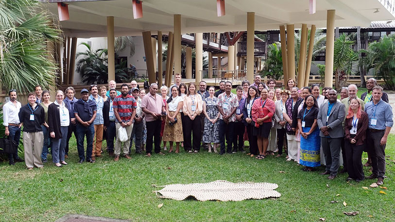 A large group of people gathered on the grass in front of a building with a palm tree off to the side and a woven mat in the foreground