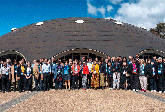 A large group of people gather outside the front of the Shine Dome on a sunny blue-sky day.