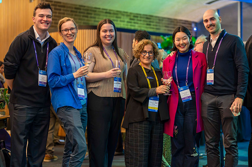 Six people pose and smile for a group photo, wearing lanyards and holding drinks