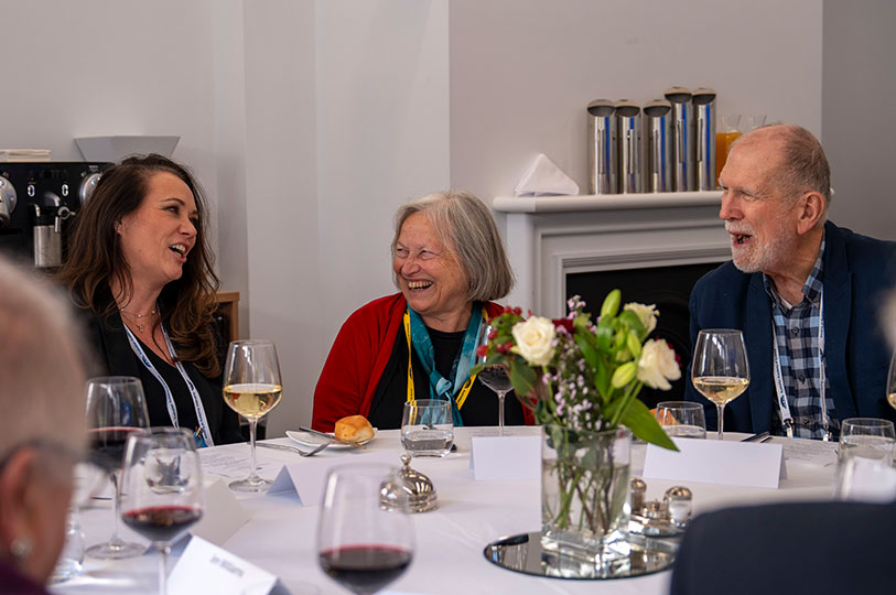 Three people sitting at a table with a white tablecloth, wine glasses and flowers, smiling and laughing