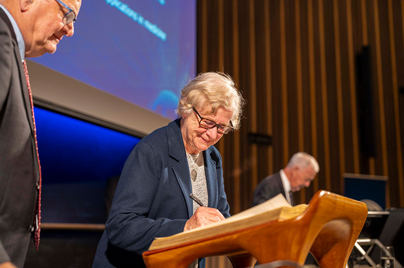 A woman signs a large book on a podium in front of a large screen