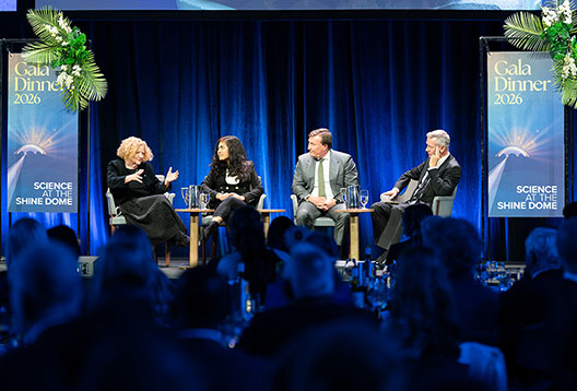 Four people sit on chairs on a stage with a blue backdrop in front of an audience