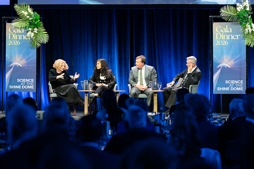 Four people sit on chairs on a stage with a blue backdrop in front of an audience.