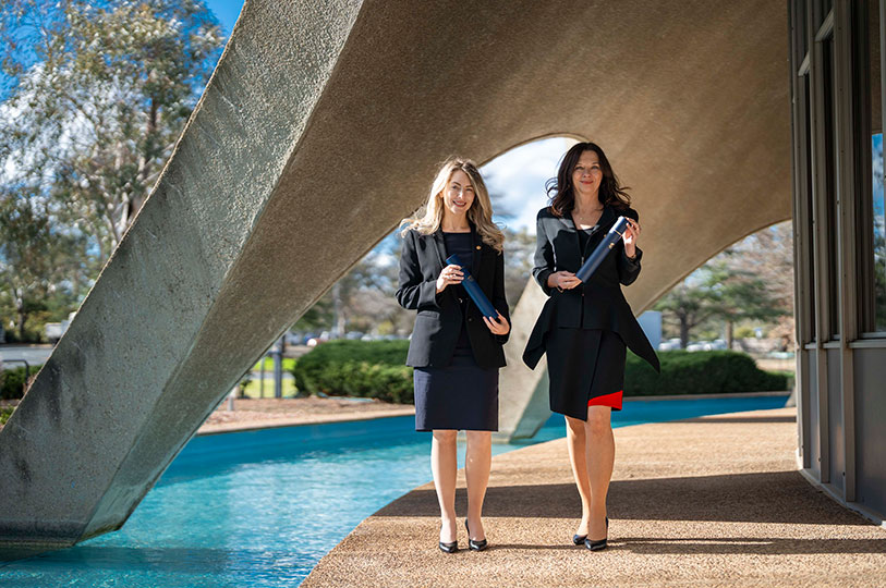 Two women in business attire hold blue certificate tubes, standing under the concrete arches of the Shine Dome, next to the moat.