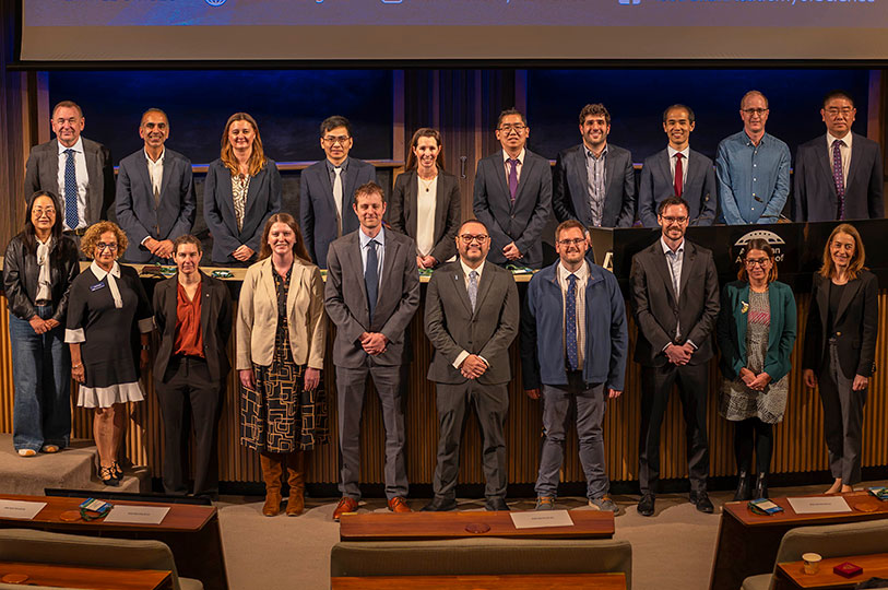 Twenty people stand in two rows for a group photo at the front of a lecture theatre