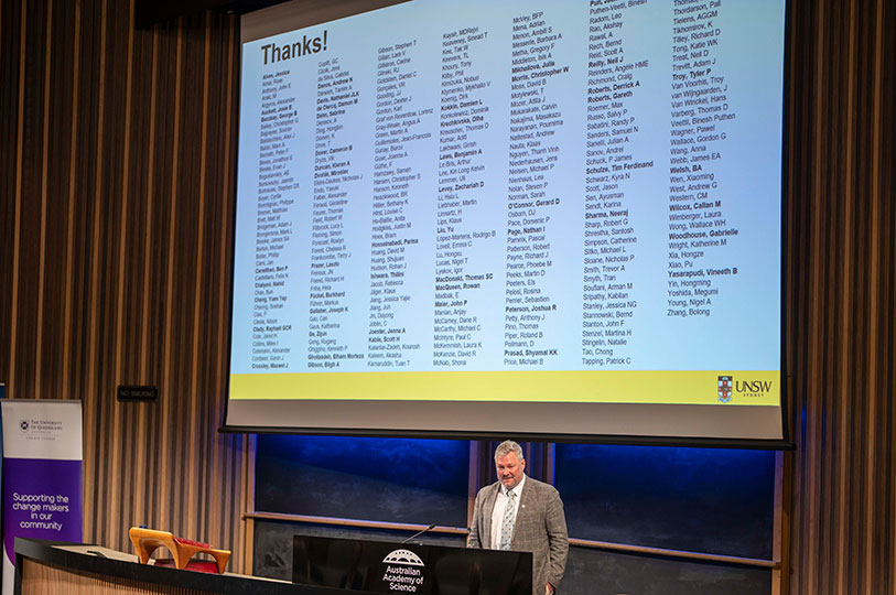 A man stands behind a lectern in front of a large projector screen displaying hundreds of names