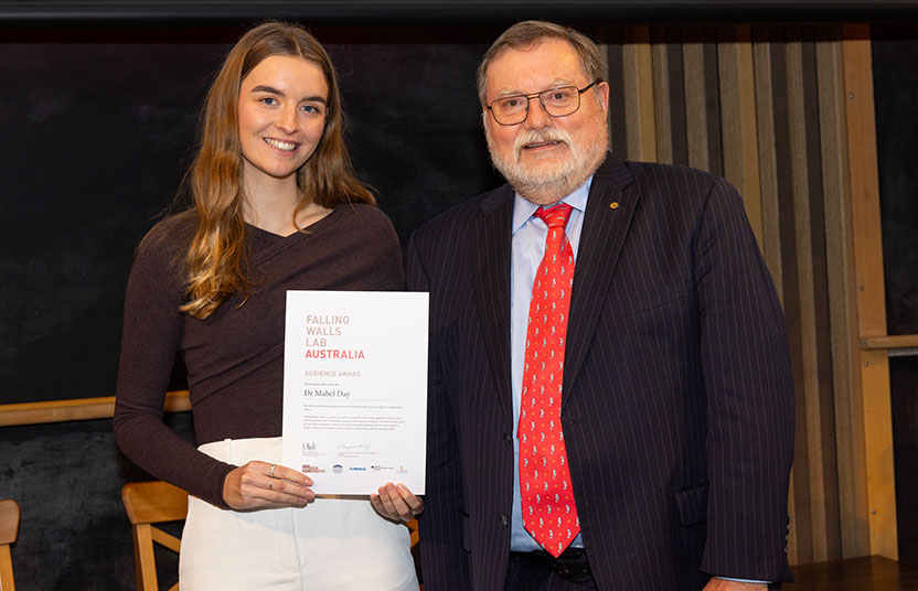 A woman on the left smiles while holding a certificate that says 'Falling Walls Lab Australia'. Next to her is a man, also smiling, wearing glasses, a suit and a red tie. They are in a room with wooden panelling on the walls.