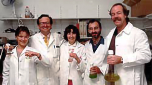 A group of two female and three male scientists in white lab coats holding lab bottles, smiling and looking relaxed