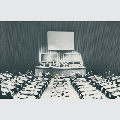 Looking from the rear to the front of a lecture theatre with seating radiating out in a semi-circular way from the stage. All seats are taken and there are people on the stage. Behind the stage are two large blackboards and above that a large white screen. Photo is black and white.
